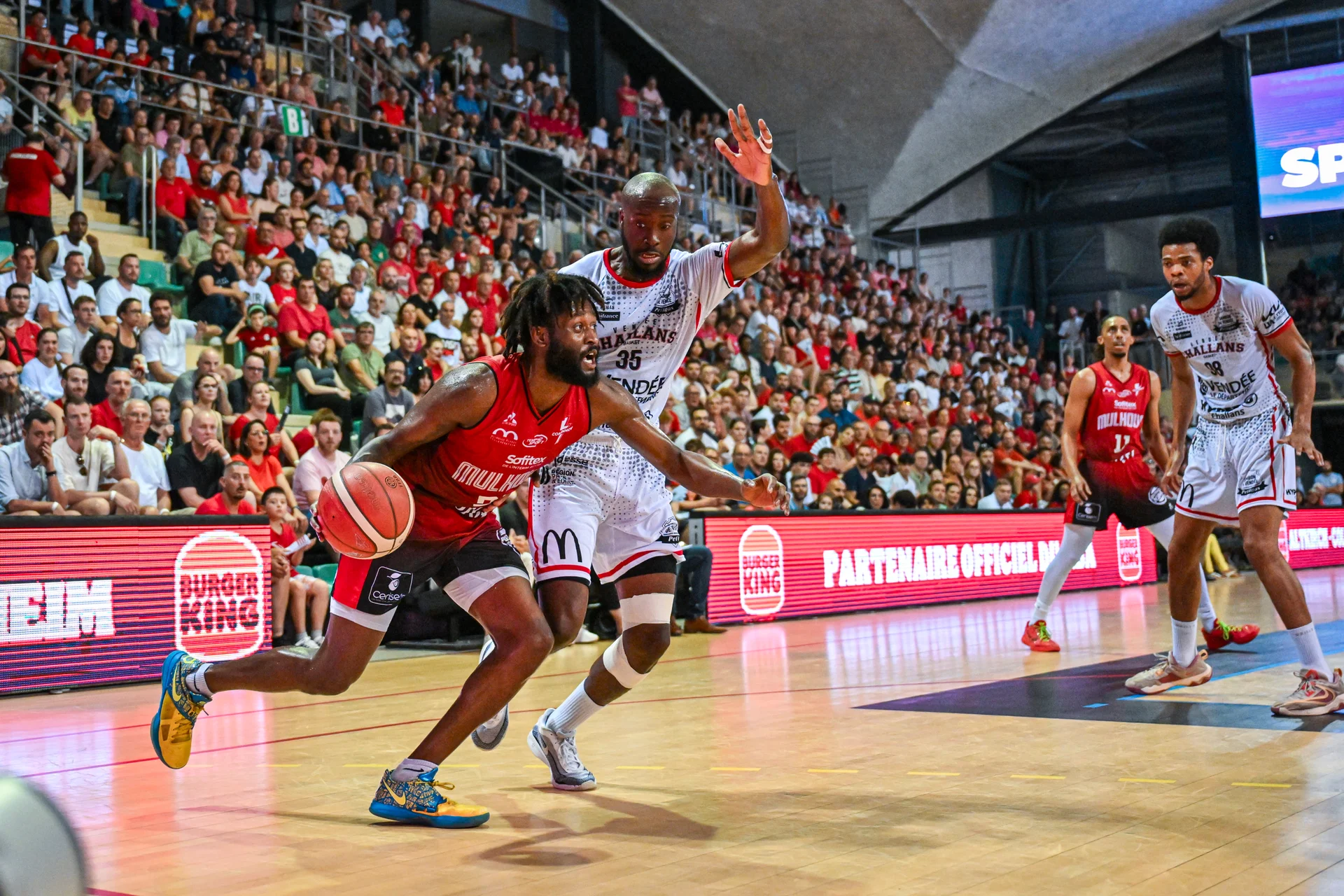 basket-pro-joueur-mulhouse-dribble-penetration Joueur de basketball de Mulhouse en pleine pénétration, défiant la défense adverse lors d'un match intense.