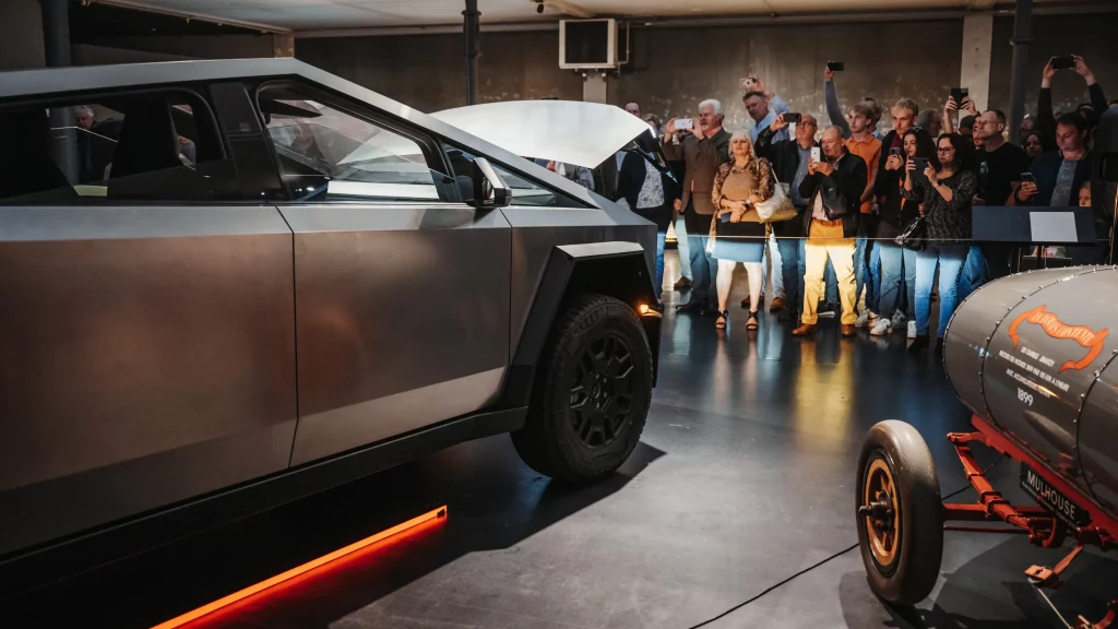 Cybertruck Tesla exposé à Mulhouse devant une foule, en contraste avec une voiture historique 1899.