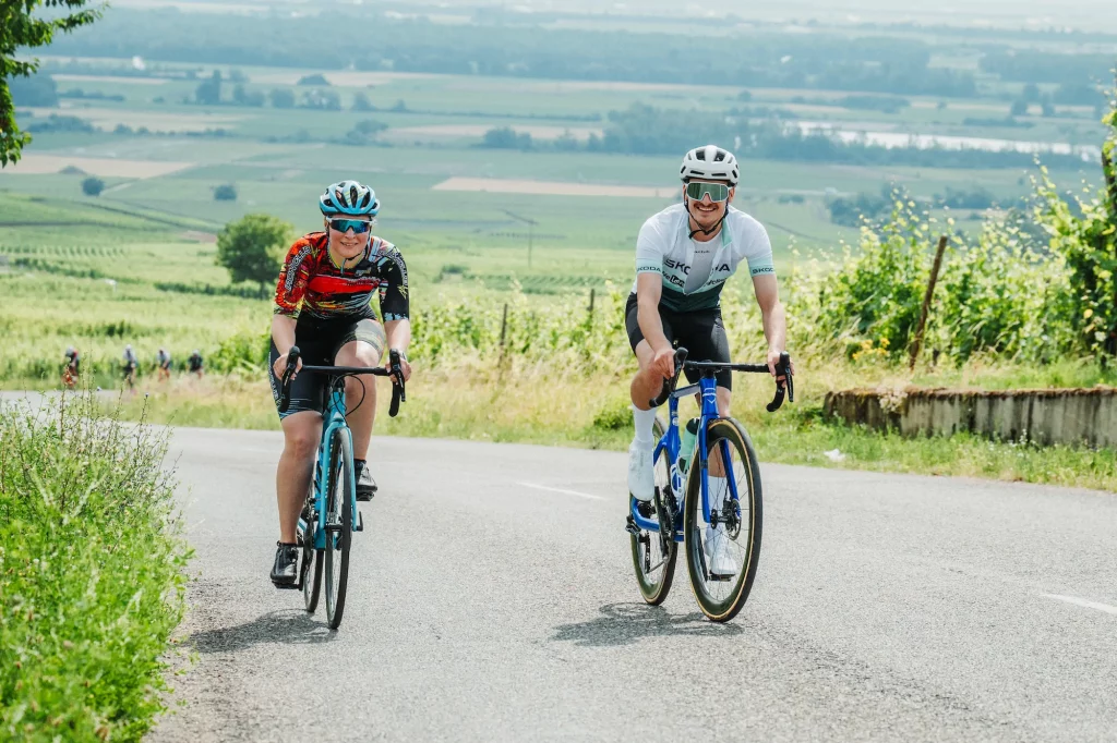 Deux cyclistes souriants grimpant une route à travers les vignobles d'Alsace pour les Boucles Škoda.