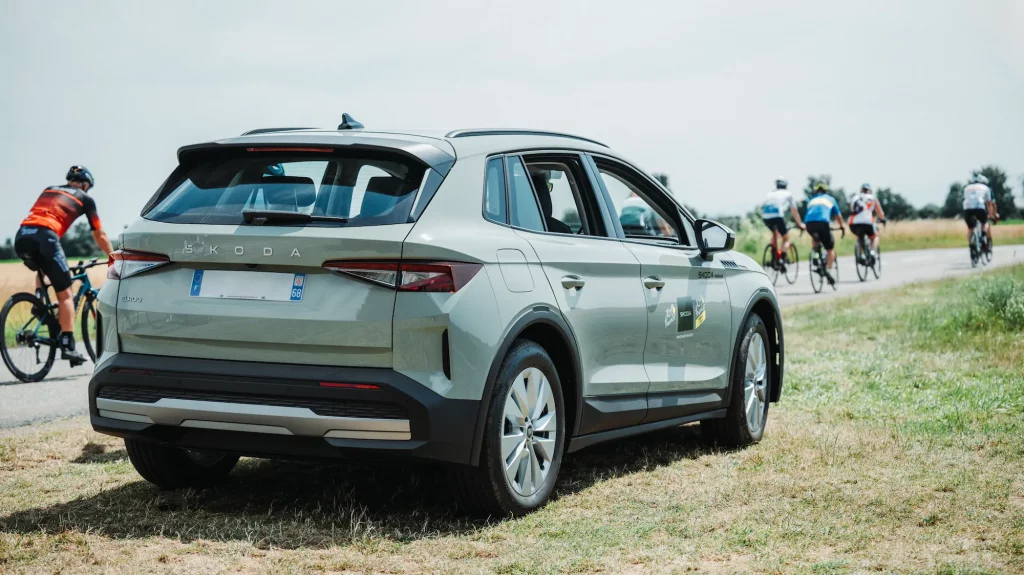 SUV Skoda Elroq stationné au bord d'une route avec des cyclistes de la boucle skoda en arrière-plan.