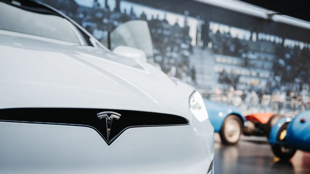 Gros plan du logo Tesla chromé sur voiture blanche, face à des voitures anciennes au Musée de Mulhouse.